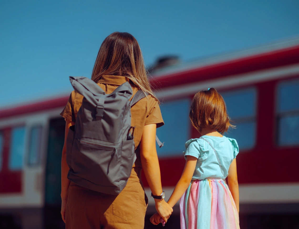 Adult and child holding hands stand in front of a moving train. The adult has a backpack, and both face the train. Sky is clear.